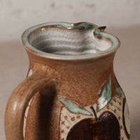 Close-up of a handmade ceramic pitcher with a brown speckled glaze, a turquoise-green rim, and painted apple motifs on the side.