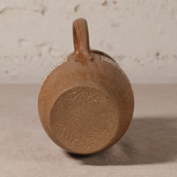 A close-up shot of the bottom of a rustic brown ceramic mug with a loop handle, resting on a neutral surface against a textured light background.