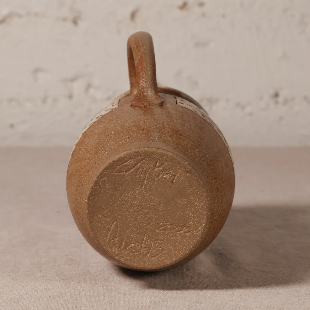 A close-up shot of the bottom of a rustic brown ceramic mug with a loop handle, resting on a neutral surface against a textured light background.