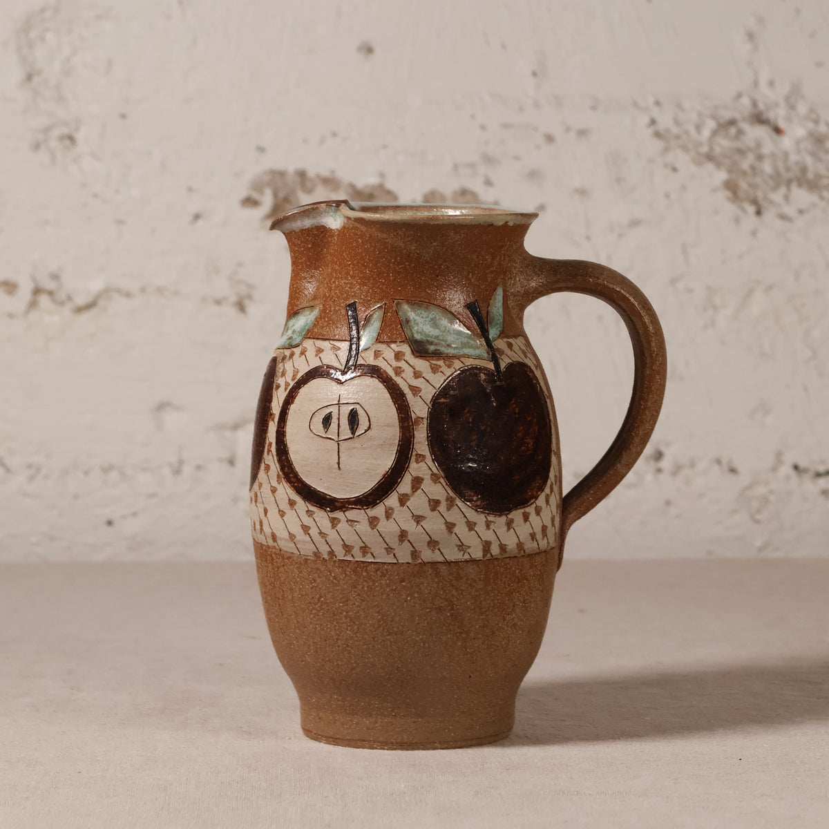 A handmade ceramic pitcher with a brown and beige speckled glaze, decorated with hand-painted dark brown apples and a patterned band, set against a rustic textured white wall.