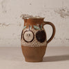 A handmade ceramic pitcher with a brown and beige speckled glaze, decorated with hand-painted dark brown apples and a patterned band, set against a rustic textured white wall.
