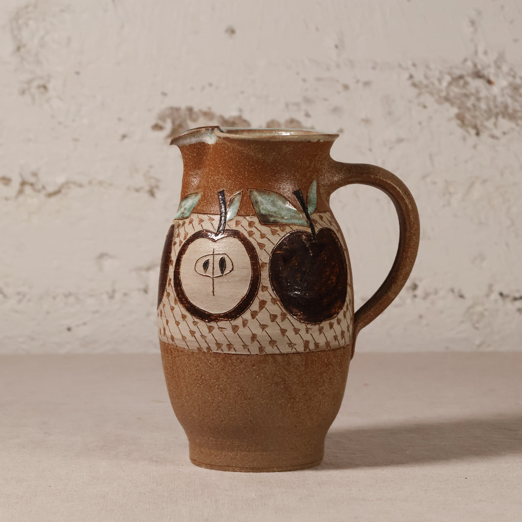A handmade ceramic pitcher with a brown and beige speckled glaze, decorated with hand-painted dark brown apples and a patterned band, set against a rustic textured white wall.