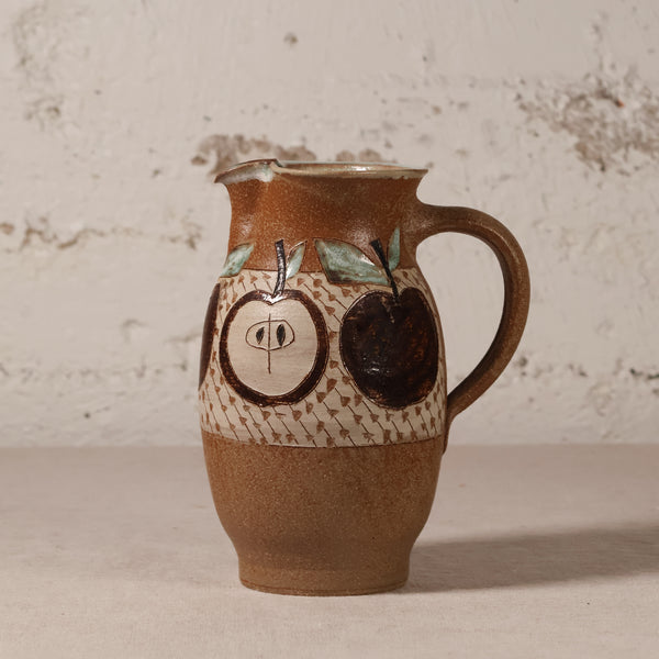 A handmade ceramic pitcher with a brown and beige speckled glaze, decorated with hand-painted dark brown apples and a patterned band, set against a rustic textured white wall.