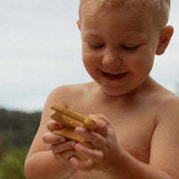 A young blonde child outdoors, holding and playing with several small yellowish sticks or pieces in their hands.