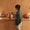 Person standing in a kitchen holding a glass, with a wooden wall and counter in the background.