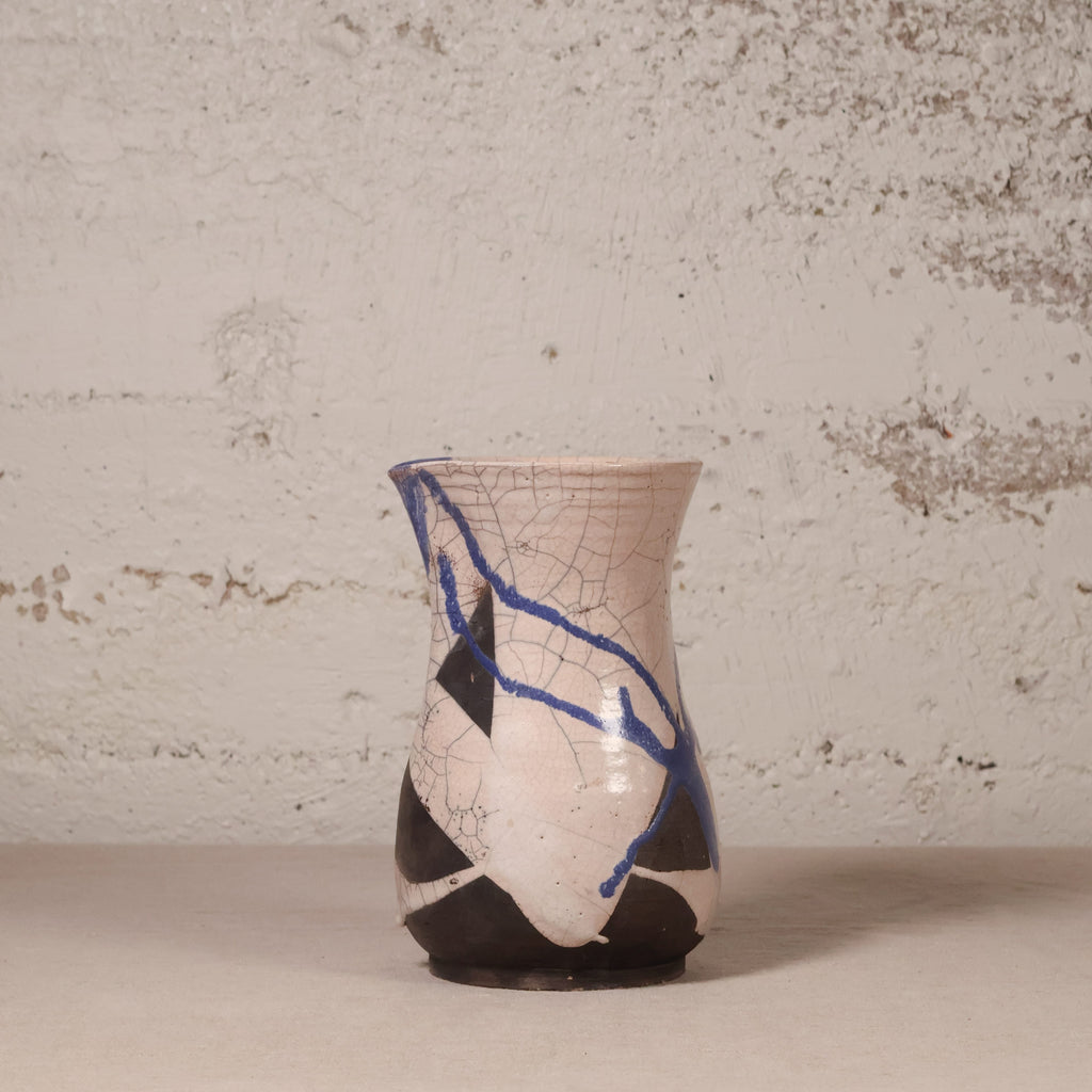 A handmade ceramic vase with a textured, hand-painted finish. The vase features a glossy off-white glaze with prominent black geometric shapes and a striking blue crackle pattern running diagonally across the surface.