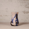 A handmade ceramic vase with a textured beige glaze, featuring abstract blue and black painted patterns and a crackled glaze finish, set against a rustic, distressed plaster wall.