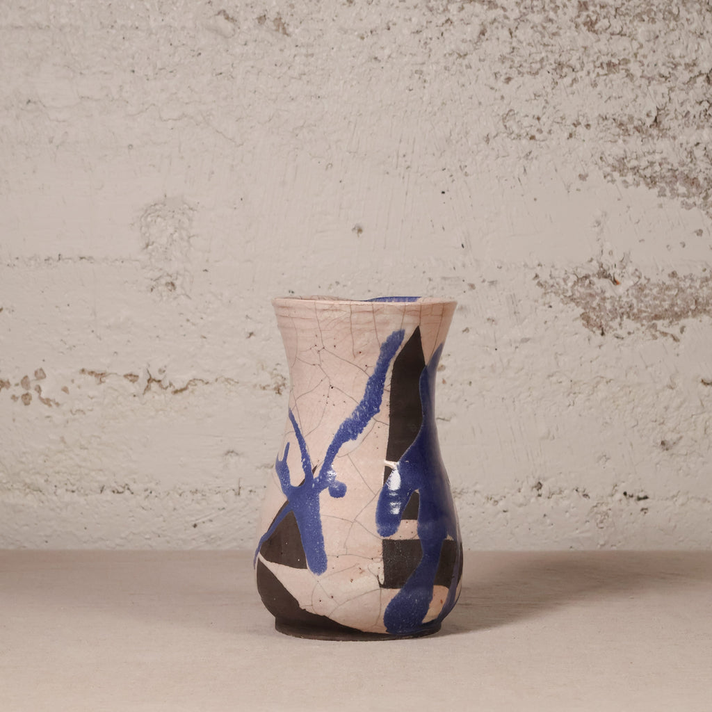 A handmade ceramic vase with a textured beige glaze, featuring abstract blue and black painted patterns and a crackled glaze finish, set against a rustic, distressed plaster wall.