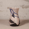 A handmade ceramic vase with a beige and black abstract pattern, decorated with blue lines and a crackle glaze, set against a rustic textured wall.
