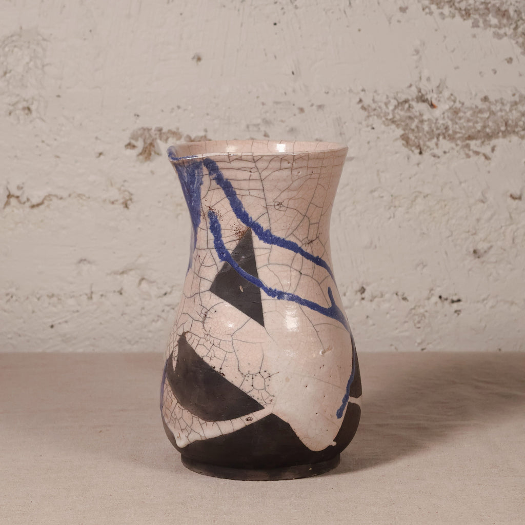 A handmade ceramic vase with a beige and black abstract pattern, decorated with blue lines and a crackle glaze, set against a rustic textured wall.