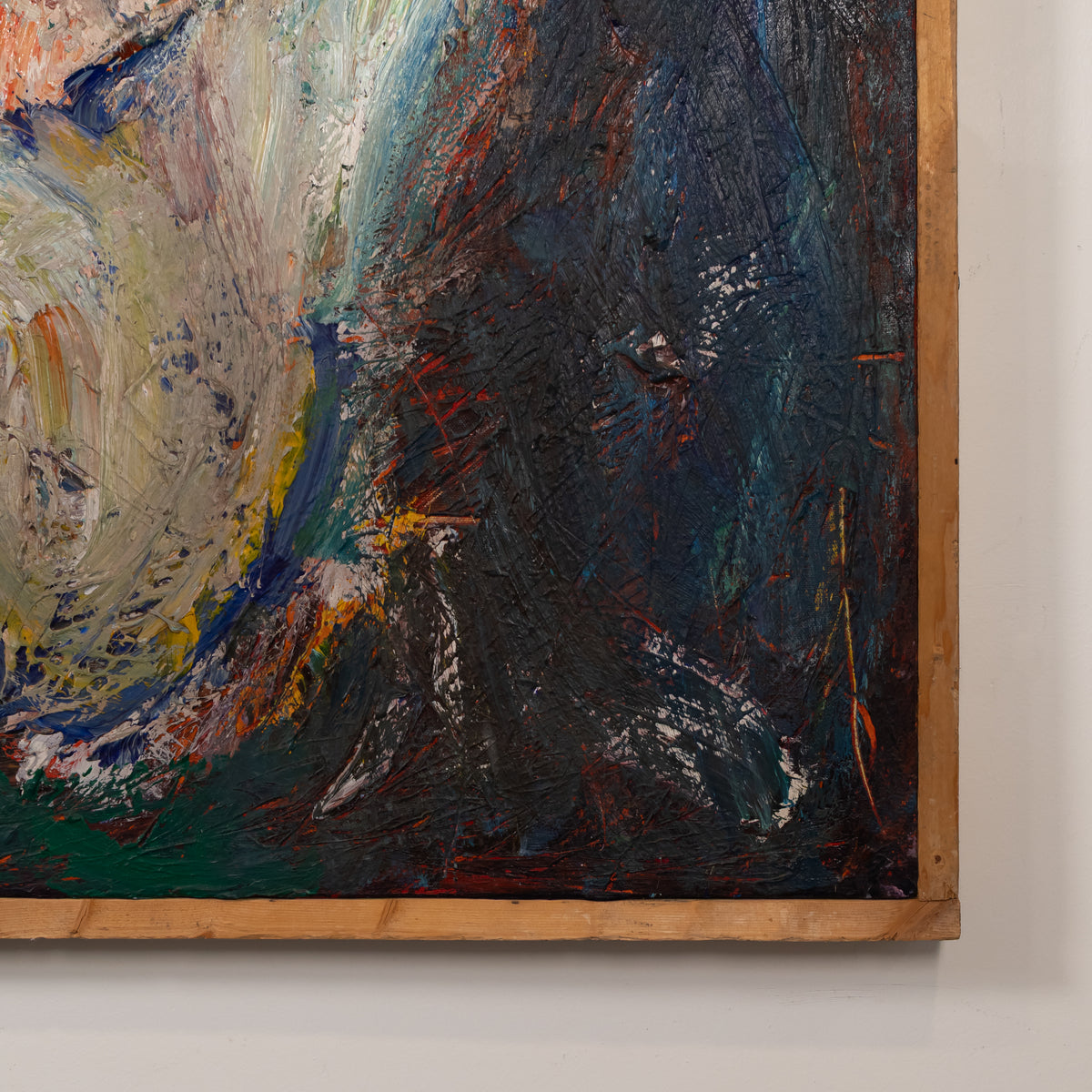 Close-up of an abstract canvas painting with thick impasto brushstrokes in dark blue, black, cream, green, and rust red tones, framed in light natural wood against a white wall.