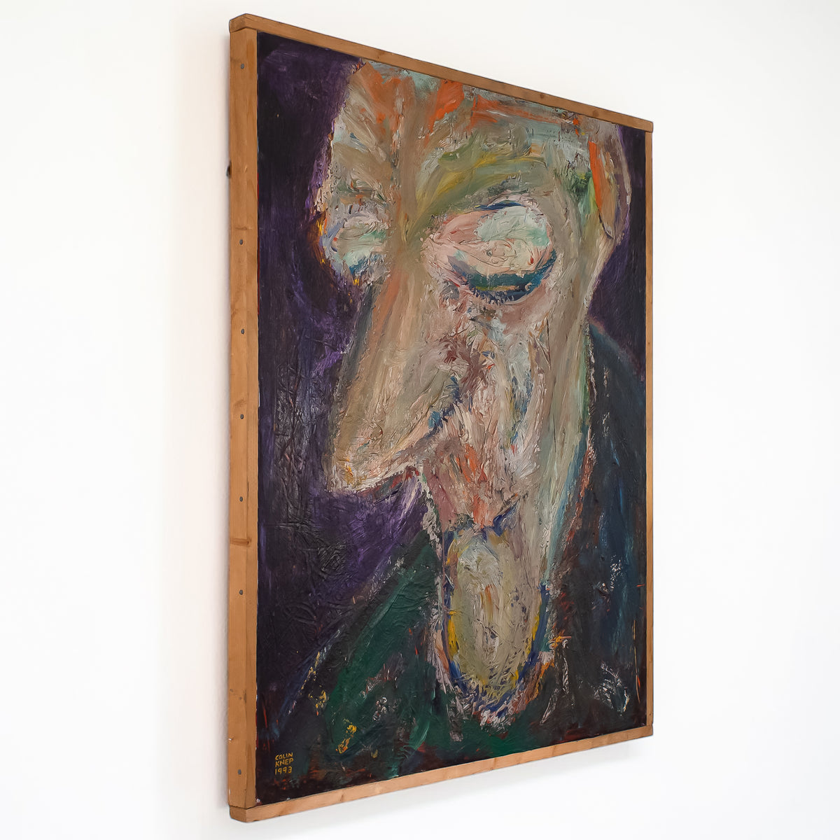An abstract portrait painting of a face with thick textured brushstrokes in purple, green, beige, orange, and blue, housed in a light brown wooden frame and hanging on a white wall.