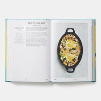 An open cookbook spread showing a recipe page with text on the left and a food photograph of eggs in a black oval skillet on the right, photographed from above on a light gray background.