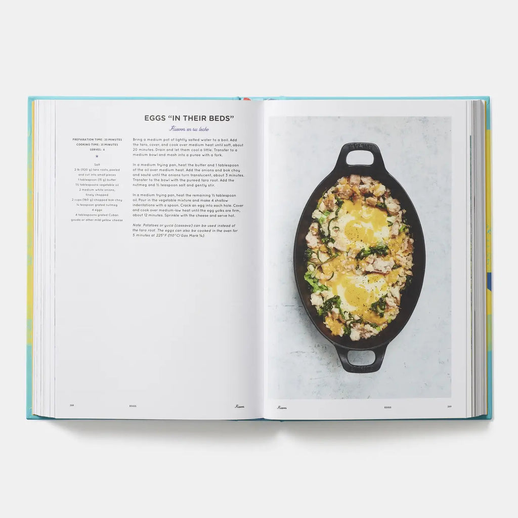 An open cookbook spread showing a recipe page with text on the left and a food photograph of eggs in a black oval skillet on the right, photographed from above on a light gray background.
