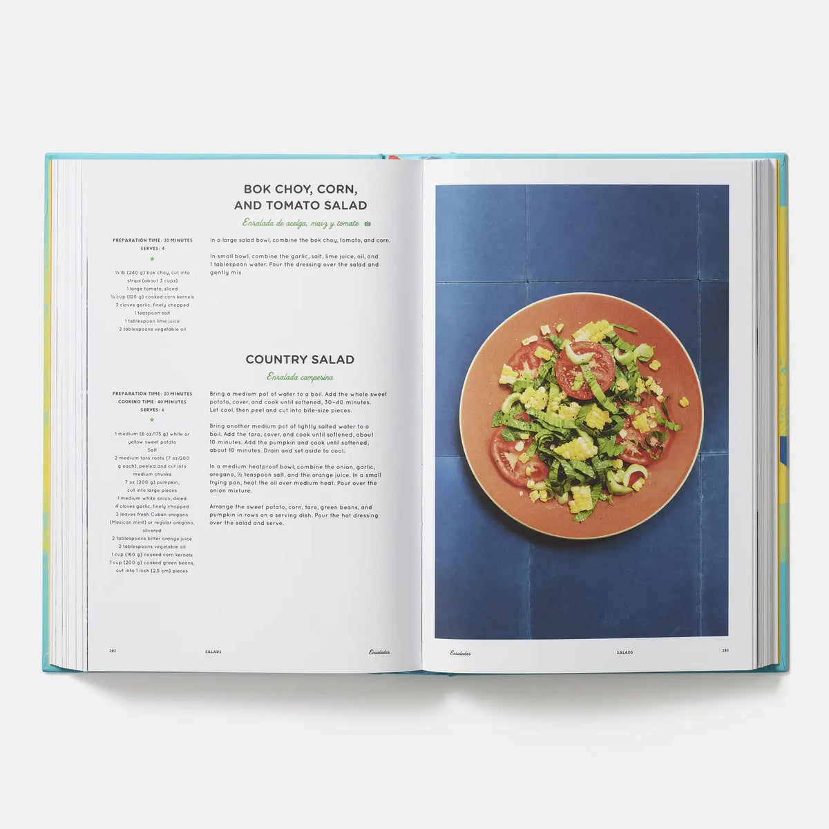 An open cookbook showing recipe pages for box choy, corn, and tomato salad and country salad, with a photo of a salad on a plate against a blue background.