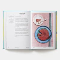An open cookbook spread showing a recipe for Yuca Flan, with printed text on the left page and a food photograph of a sliced red-pink dessert on a blue plate on the right page, photographed on a light gray background.
