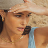 Woman shielding her eyes from the sun with a beige hat, wearing gold jewelry.