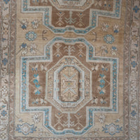 A close-up of a traditional patterned rug with a central medallion, featuring a beige and brown base with light blue and cream accents.