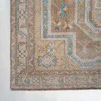 A close-up of a traditional patterned rug corner showing beige, tan, cream, and light blue motifs with a woven texture.