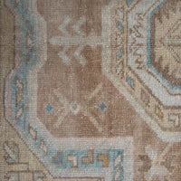 Close-up of a woven rug showing a beige and tan field with geometric motifs in light blue, cream, and small yellow accents.
