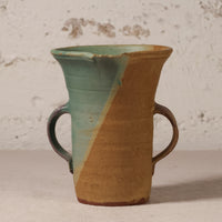 A handmade ceramic pitcher with two side handles, featuring a diagonal split glaze of light teal green and warm tan, set against a textured off-white wall.