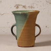 A handmade ceramic pitcher with two side handles, featuring a diagonal split glaze of light teal green and warm tan, set against a textured off-white wall.