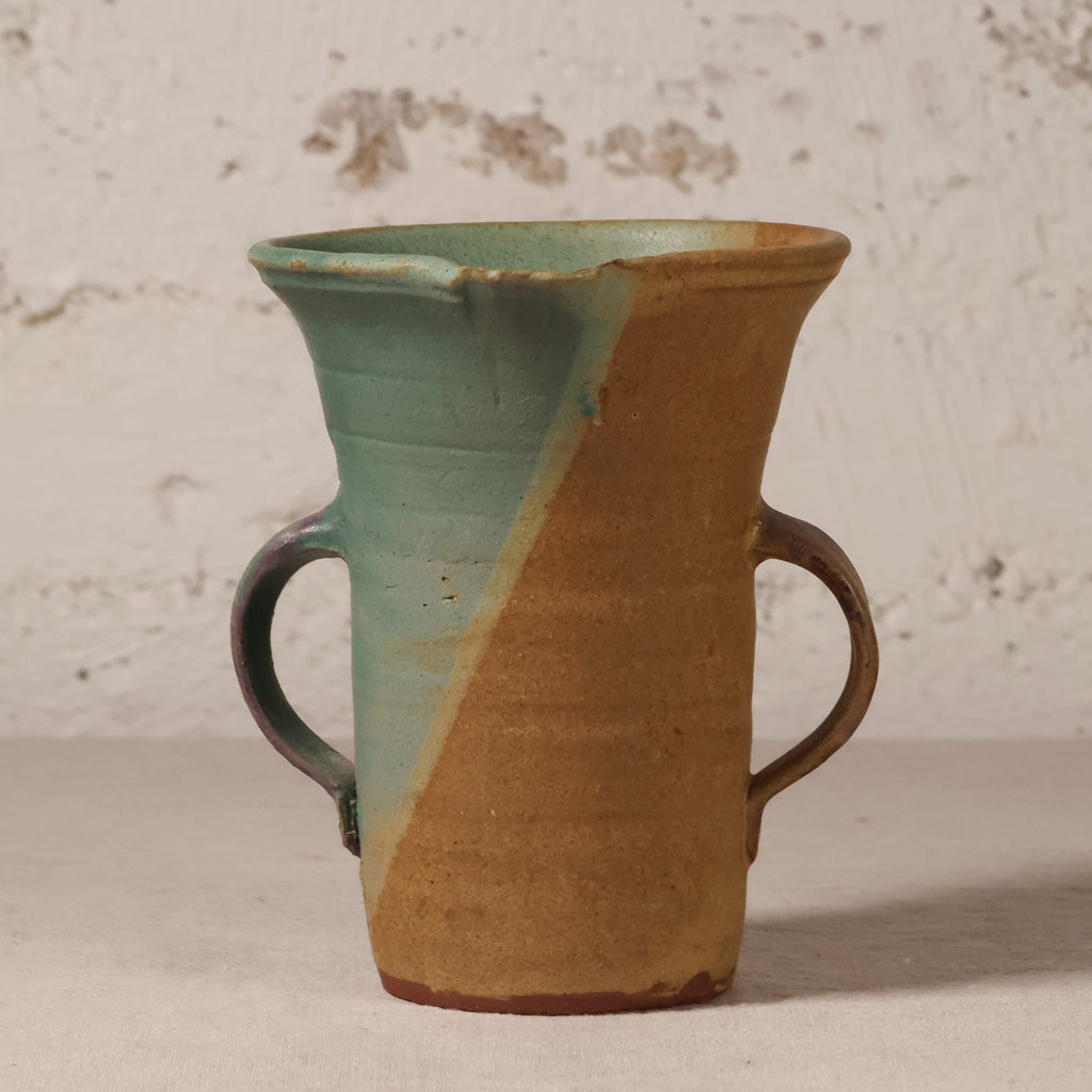 A handmade ceramic pitcher with two side handles, featuring a diagonal split glaze of light teal green and warm tan, set against a textured off-white wall.