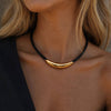A close-up of a model wearing a black cord necklace with a polished gold crescent-shaped pendant.