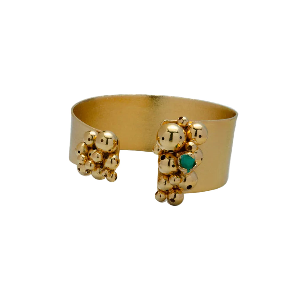 A gold-toned wide cuff bracelet with textured, beaded clusters on both ends and a small green stone accent, shown on a light gray background.