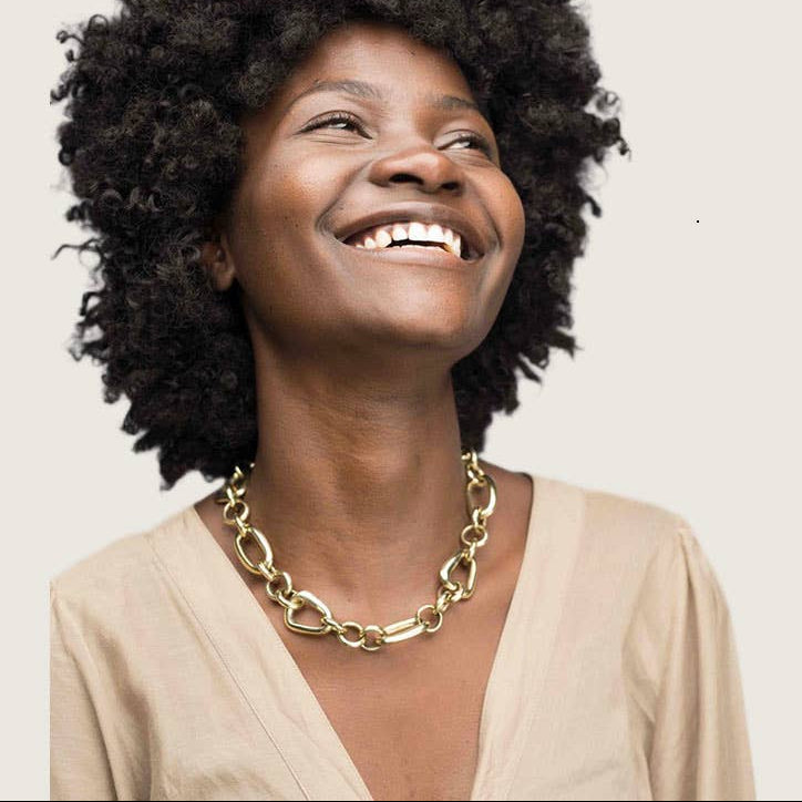 A smiling woman with a voluminous black afro hairstyle wearing a chunky gold chain necklace and a beige top, photographed against a solid light beige background.