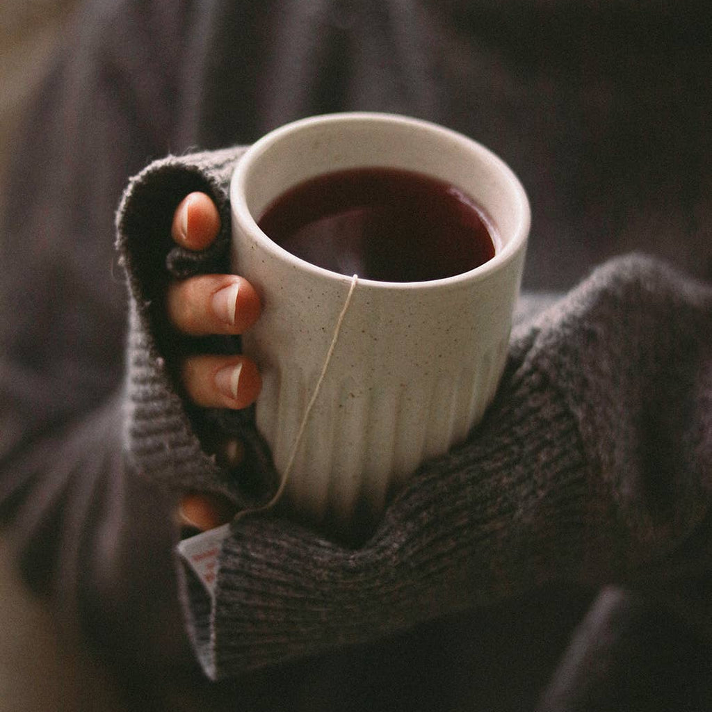 A close-up lifestyle shot of a person wearing a dark gray knit sweater and fingerless gloves, holding a white ceramic mug filled with a dark tea or coffee. A tea bag string hangs from the rim of the mug.