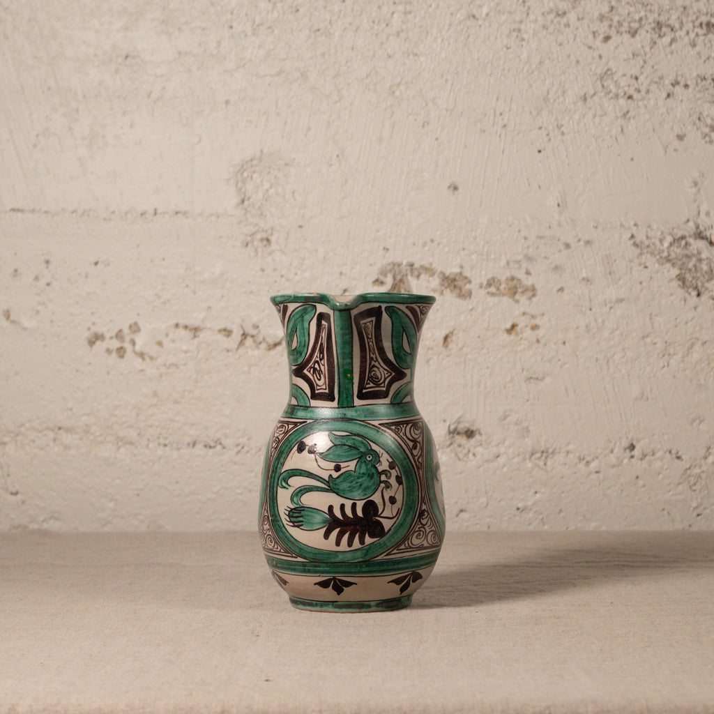 Ceramic pitcher with green and black floral patterns on a textured surface.