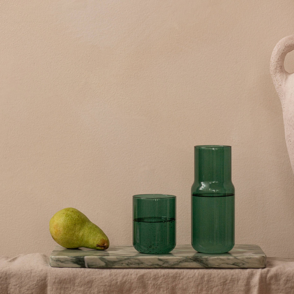 Two translucent dark green glass vessels, one short tumbler and one taller bottle, displayed on a marble tray against a warm beige wall, with a pear and a large textured vase as props.