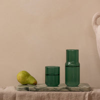 Two translucent dark green glass vessels, one short tumbler and one taller bottle, displayed on a marble tray against a warm beige wall, with a pear and a large textured vase as props.