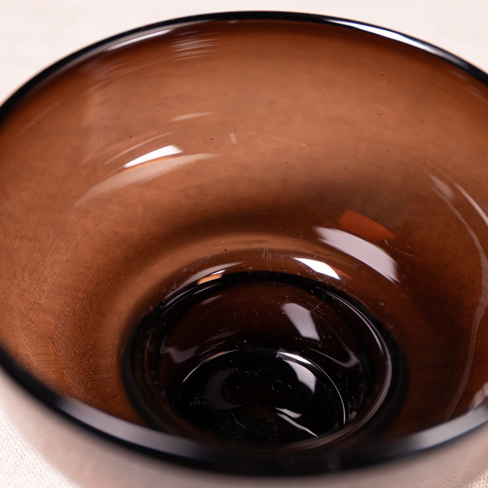 A close-up of a translucent amber-brown glass bowl with a glossy finish and a dark rim, photographed against a light neutral background.