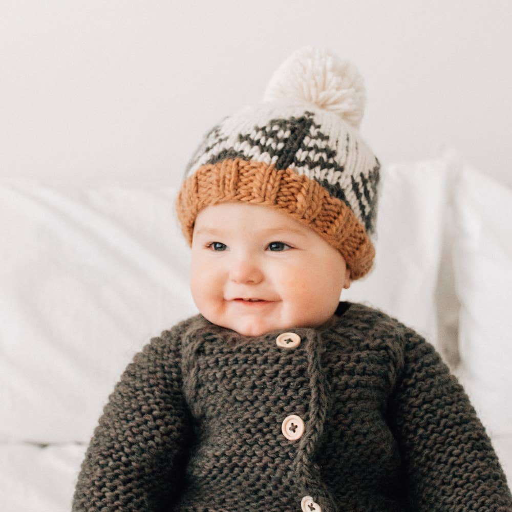 A baby wearing a chunky knit pom-pom hat in cream, brown, and gray, paired with a matching dark gray knit cardigan, sitting against a soft white background.