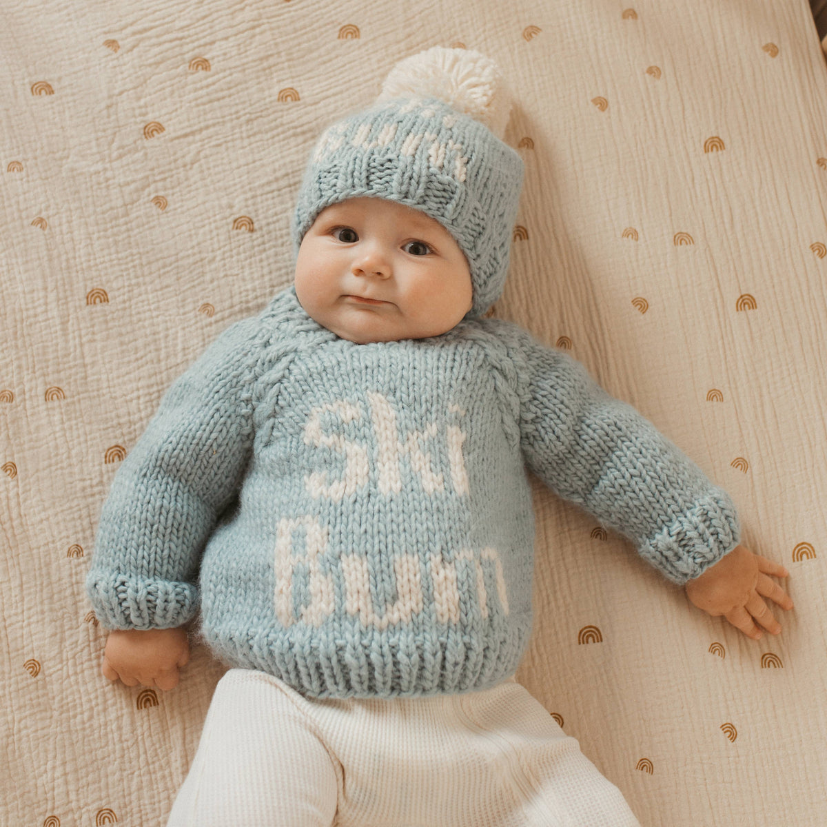 A baby lying on a cream muslin blanket with small gold rainbows, wearing a light blue hand-knit sweater and matching beanie hat with white yarn lettering that reads 'Ski Bum'.