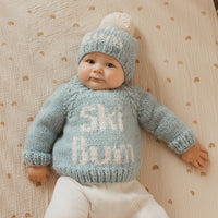 A baby lying on a cream muslin blanket with small gold rainbows, wearing a light blue hand-knit sweater and matching beanie hat with white yarn lettering that reads 'Ski Bum'.