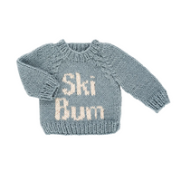 A hand-knit baby sweater in a dusty slate blue color, featuring the words 'Ski Bum' knitted in cream-colored yarn across the chest. The sweater is laid flat on a clean white background.