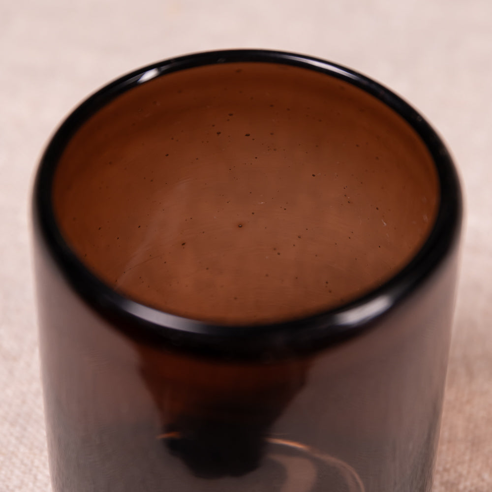 A close-up of a dark amber brown glass tumbler with a glossy black rim, set against a neutral beige textured background.