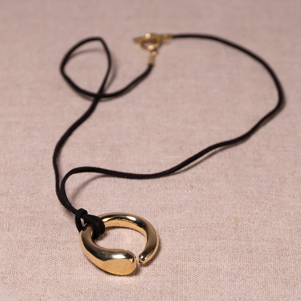 A gold-toned metal pendant necklace with a black cord, displayed on a textured beige fabric background.