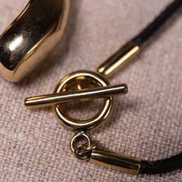 Close-up of a gold-tone toggle ring clasp with a black cord, resting on a textured beige fabric surface.