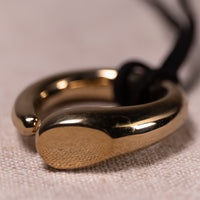 A close-up of a polished gold-toned ring with a textured front face and a black cord attached, resting on a light beige fabric surface.