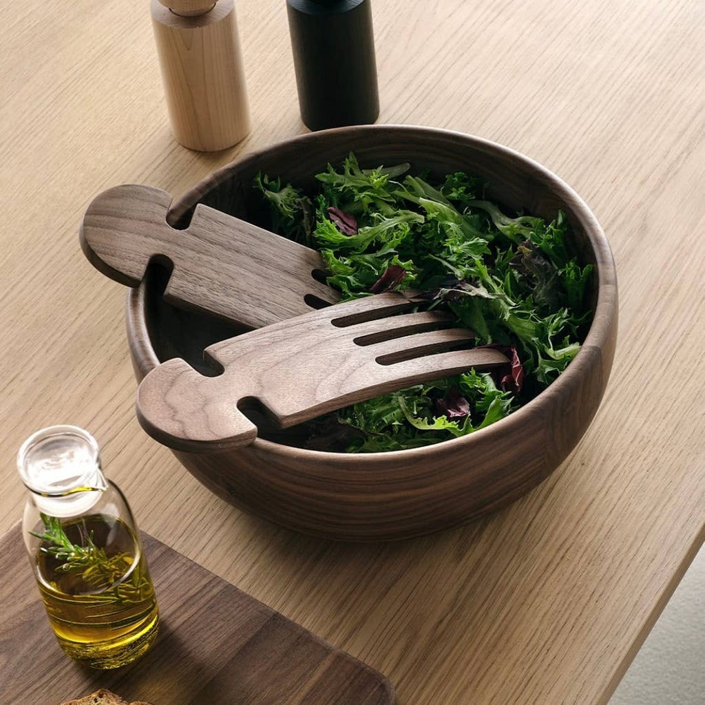 Wooden salad bowl with serving utensils and a small bottle of oil on a wooden table.