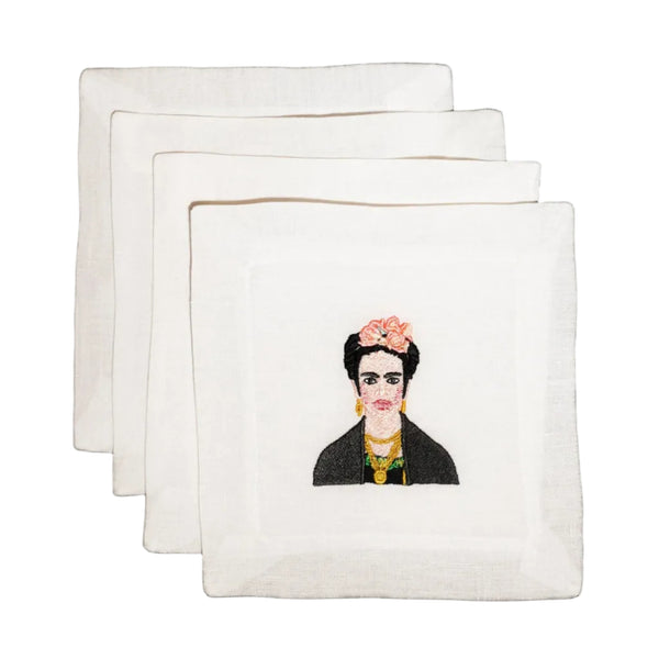 Three square white embroidered napkins are stacked in a fan arrangement on a white background. The front napkin features an embroidered portrait of Frida Kahlo with black hair, a pink flower crown, and a black jacket.