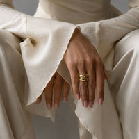 A close-up of a person wearing a cream-colored textured dress with dramatic flared sleeves, seated against a neutral gray background. The person is wearing a gold ring with three interlocking curved bands on one finger.