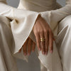 A close-up of a person wearing a cream-colored textured dress with dramatic flared sleeves, seated against a neutral gray background. The person is wearing a gold ring with three interlocking curved bands on one finger.