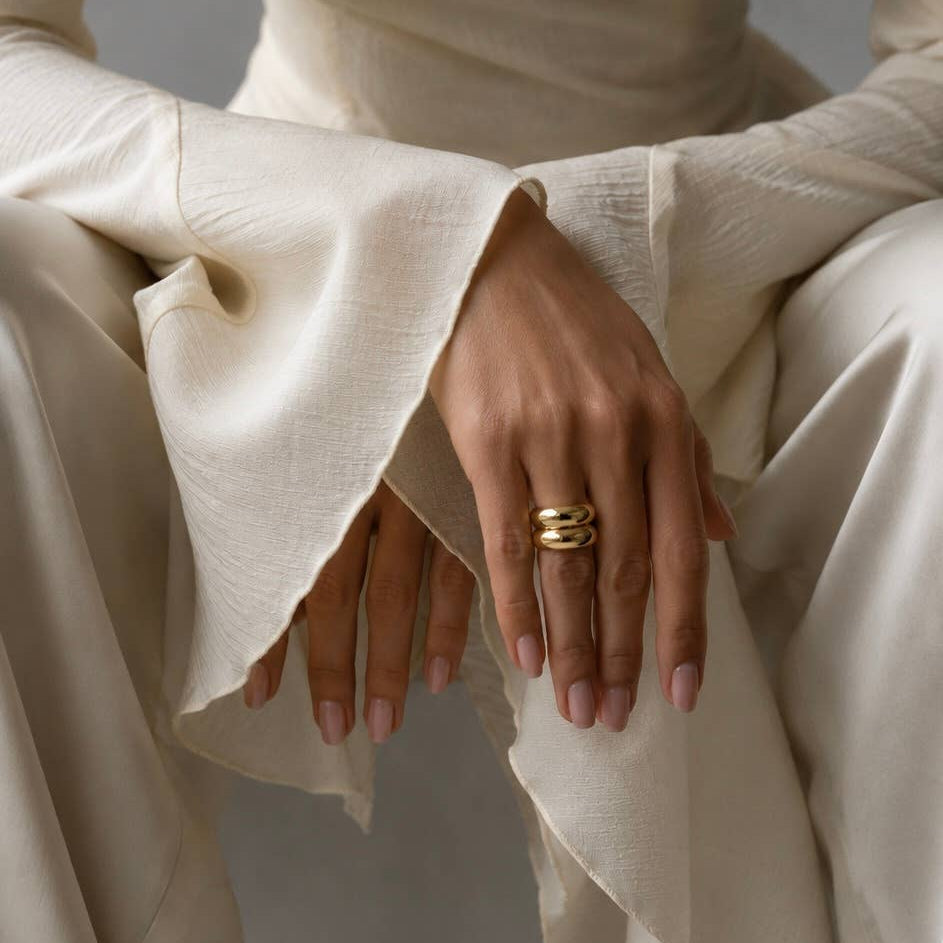 A close-up of a person wearing a cream-colored textured dress with dramatic flared sleeves, seated against a neutral gray background. The person is wearing a gold ring with three interlocking curved bands on one finger.