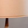 Close-up of a table lamp shade in a warm beige or peach-toned fabric, with part of the dark brown lamp base visible against a plain gray background.