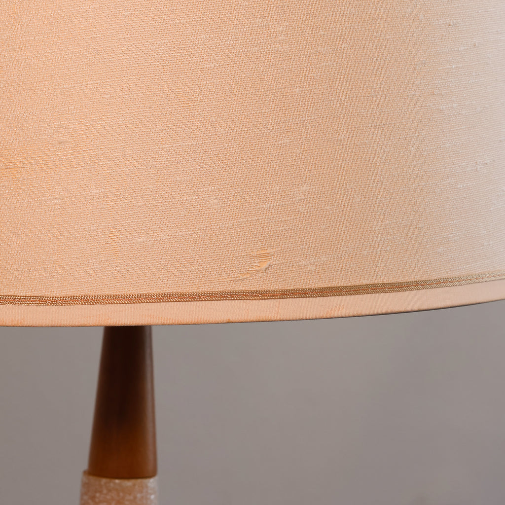Close-up of a table lamp shade in a warm beige or peach-toned fabric, with part of the dark brown lamp base visible against a plain gray background.
